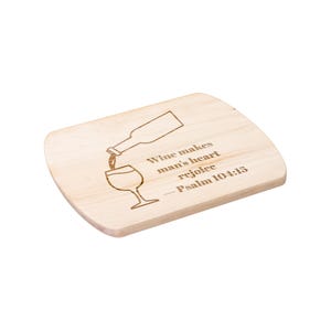 May include: A light-coloured wooden chopping board with rounded edges. The board features an etched design of a wine bottle pouring into a wine glass, with the text "Wine makes man's heart rejoice - Psalm 104:15."
