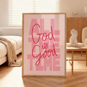 Op de afbeelding: Een ingelijste roze kunstprint met de tekst "ALL GOD IS GOOD ALL THE TIME" in een mix van vette en scriptlettertypen. Het kunstwerk heeft een lichtroze achtergrond en een houten frame.