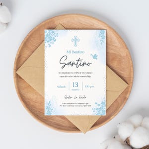Puede incluir: Una invitación de bautizo con el texto "Mi Bautizo Santino" en escritura elegante, con una cruz y una ilustración de paloma. La invitación es azul claro y blanca, con detalles florales. Incluye la fecha, hora y detalles del lugar, sobre una bandeja de madera.