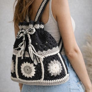 Może przedstawiać: Ręcznie robiony plecak z szydełka w kolorach czarnym, szarym i kremowym. Torba posiada zapięcie na sznurek z frędzlami i kwiatowy wzór. Paski również są szydełkowane, w stylu boho.