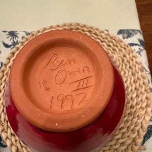May include: A red ceramic bowl with a terracotta base, on a woven placemat. The base is inscribed with "Ben Owen" and the year "1992". The bowl's interior is a glossy red. The bowl is likely handmade.