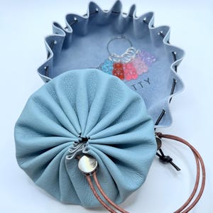 May include: A light blue leather drawstring pouch with a brown cord and silver button closure. A matching blue leather tray with a scalloped edge and a keychain with colorful gummy bear charms are also visible.