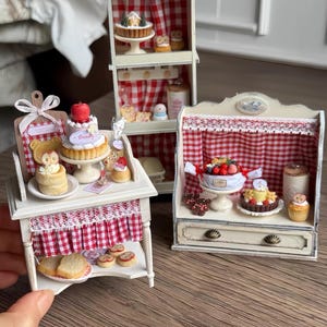 Puede incluir: Miniatura de exhibición de panadería con pasteles y pasteles. Las exhibiciones son blancas con cortinas de cuadros rojos y blancos y un techo de madera. Los pasteles en miniatura incluyen un pastel con una manzana roja encima.