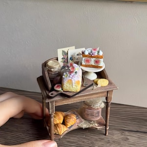 Puede incluir: Mesa de madera en miniatura con una selección de productos horneados. Incluye cruasanes, una rebanada de pastel con higos y un panecillo cubierto con flores. También hay tarros de conservas y tarjetas decorativas. La escena está ambientada en una panadería en miniatura.
