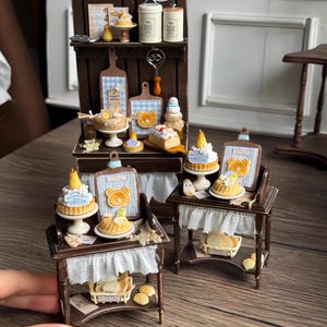 Puede incluir: Miniatura de exhibición de panadería con pasteles, pasteles y tablas de cortar. La exhibición presenta una estructura de madera marrón oscuro con estantes, un borde de encaje blanco y varios productos horneados. Los pasteles en miniatura tienen glaseado amarillo y blanco.