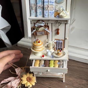 Puede incluir: Miniatura de gabinete blanco con estantes, cajones y varios objetos decorativos. Incluye libros en miniatura, una cesta, un pastel, pasteles y una figura de cisne. El gabinete está pintado de blanco con detalles en azul.