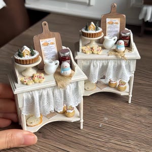 Puede incluir: Miniatura de exhibición de panadería blanca con una cortina de encaje, con pequeños pasteles, galletas y una figura de muñeco de nieve. Cada exhibición tiene una tabla de cortar de madera con un menú. Los alimentos en miniatura están en una mesa blanca.