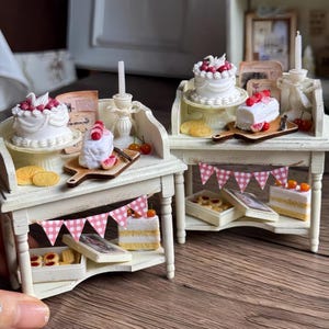 Puede incluir: Pequeñas mesas blancas de exhibición de panadería con pasteles, galletas y pasteles. Cada mesa tiene un pequeño soporte para pasteles, una tabla de cortar de madera con una rebanada de pastel y una pancarta rosa y blanca a cuadros. Velas y cerezas.