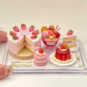 Peut inclure: Miniatures de desserts sur un plateau transparent. Un gâteau rose aux fraises, une coupe glacée, un macaron, une part de gâteau et un dessert à la gelée rouge sont disposés. Des ustensiles dorés sont également présents. Le texte "KITTY BAKERY" et "Cake" sont visibles.