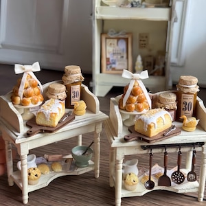 Puede incluir: Escena de cocina en miniatura con dos mesas blancas que exhiben productos horneados, frascos y utensilios. Cada mesa tiene un pastel, una tabla de cortar de madera y un cuenco pequeño. La escena incluye un armario con objetos en miniatura.