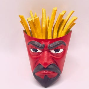 May include: A red, devil-faced container filled with yellow french fries. The container has a stern expression with black eyebrows, a black mustache, and a goatee. The fries are upright, giving the appearance of hair.