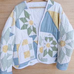 May include: A quilted jacket featuring a patchwork design with floral and gingham patterns in shades of sage green, pale yellow, and light blue on a white background. The jacket has a V-neckline, long sleeves, and two front patch pockets.
