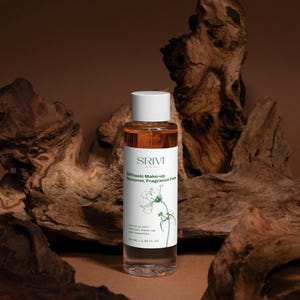 May include: A clear glass bottle of SRIVI Biphasic Make-up Remover, Fragrance Free, with a white cap. The bottle contains a clear and amber liquid. The label features the brand name and a botanical illustration. The background includes dark brown wood.