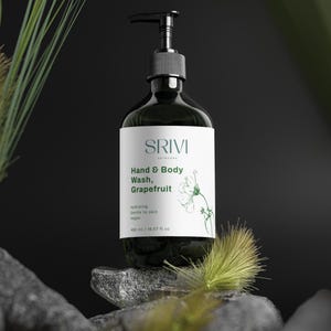 May include: A dark green hand and body wash bottle with a black pump dispenser. The label reads "SRIVI Skincare Hand & Body Wash, Grapefruit." The bottle is on a stone surface with greenery in the background. The bottle contains 490 ml / 16.57 fl oz.