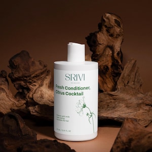 May include: A white bottle of SRIVI Fresh Conditioner, Citrus Cocktail, with a flip-top cap. The label includes text and a floral illustration. The bottle is set against a backdrop of dark wood and a brown background. The bottle contains 12.51 fl oz.