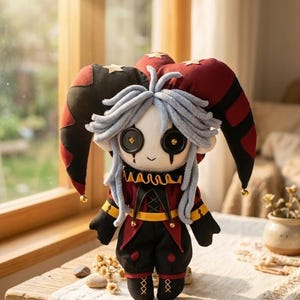 May include: A handmade plush doll in a jester costume. The doll has a white face with large button eyes, gray yarn hair, and a small smile. It wears a red and black outfit with gold accents and a matching hat with bells.