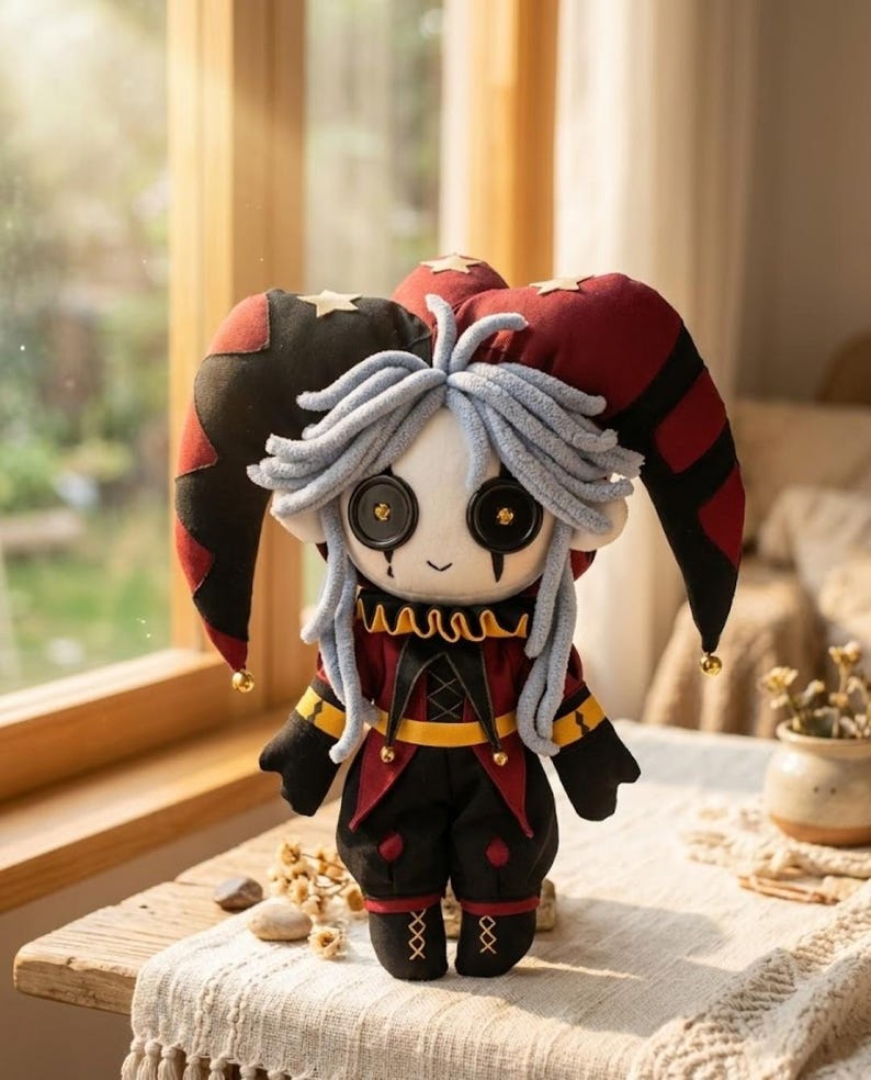 May include: A handmade plush doll in a jester costume. The doll has a white face with large button eyes, gray yarn hair, and a small smile. It wears a red and black outfit with gold accents and a matching hat with bells.