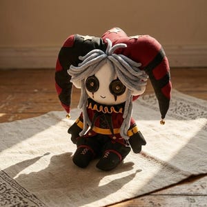 May include: A plush jester doll with a red, black, and gold color scheme. The doll has a white face with button eyes, gray hair, and a red and black jester hat. The doll is sitting on a patterned rug.