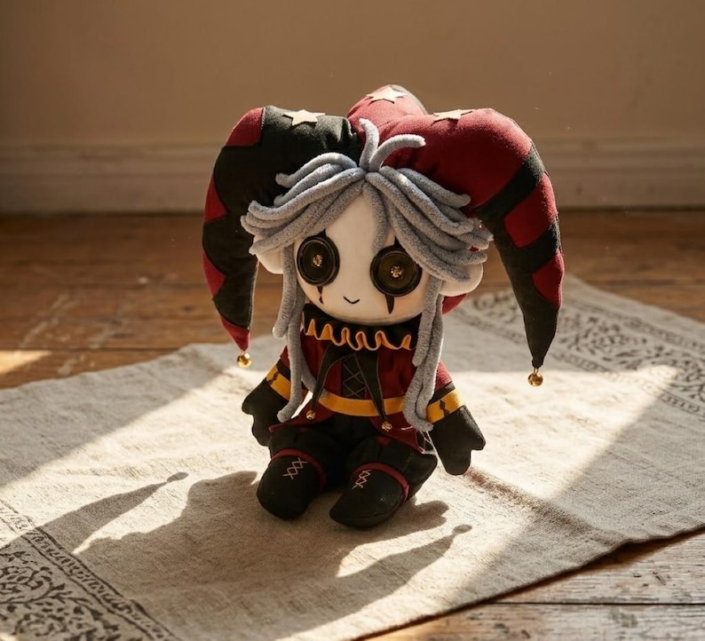 May include: A plush jester doll with a red, black, and gold color scheme. The doll has a white face with button eyes, gray hair, and a red and black jester hat. The doll is sitting on a patterned rug.