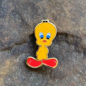 Pu&ograve; includere: Spilla smaltata gialla di Tweety Bird con bordo dorato. La spilla raffigura il personaggio dei cartoni animati con occhi blu, becco arancione e piedi rossi. La spilla &egrave; posta su uno sfondo grigio scuro strutturato.