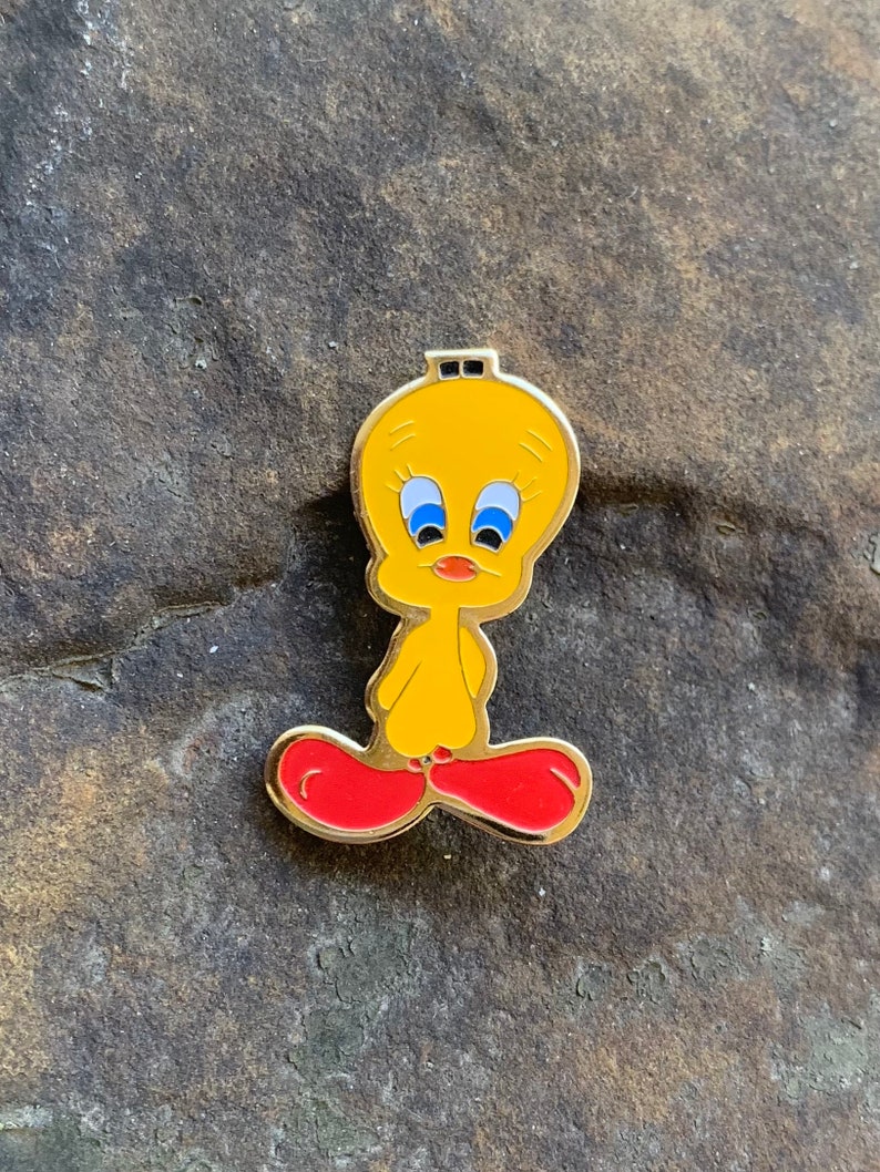 Pu&ograve; includere: Spilla smaltata gialla di Tweety Bird con bordo dorato. La spilla raffigura il personaggio dei cartoni animati con occhi blu, becco arancione e piedi rossi. La spilla &egrave; posta su uno sfondo grigio scuro strutturato.