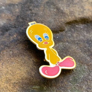 Pu&ograve; includere: Una spilla in smalto giallo raffigurante il personaggio dei cartoni animati Titti. La spilla ha un contorno dorato, occhi blu e piedi rosa. La spilla &egrave; posta su uno sfondo marrone strutturato.