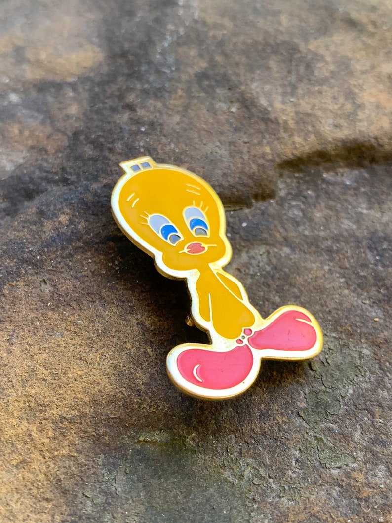 Pu&ograve; includere: Una spilla in smalto giallo raffigurante il personaggio dei cartoni animati Titti. La spilla ha un contorno dorato, occhi blu e piedi rosa. La spilla &egrave; posta su uno sfondo marrone strutturato.