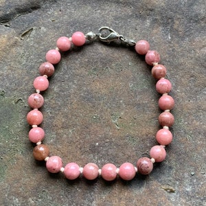 Può includere: Un braccialetto di perline rosa con chiusura argentata. Il braccialetto presenta perline rotonde e lucide in varie tonalità di rosa e marrone, infilate su un cordoncino bianco. La chiusura è un design semplice e sicuro.