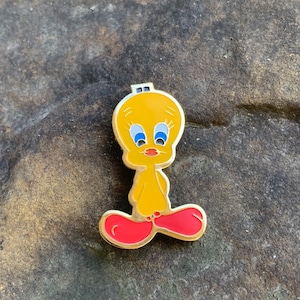 Pu&ograve; includere: Una spilla in smalto giallo raffigurante il personaggio dei cartoni animati Titti. La spilla ha un contorno dorato, occhi blu e piedi rossi. La spilla &egrave; posta su una superficie di pietra grigia strutturata.
