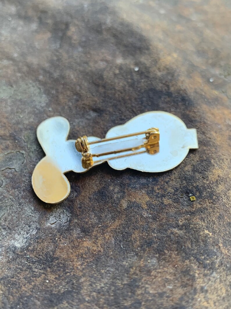 Pu&ograve; includere: Una spilla in stile vintage con un design bianco e oro. La spilla presenta una forma stilizzata con un retro dorato e una chiusura a spilla da balia. La spilla &egrave; probabilmente in metallo e ha una finitura lucida.
