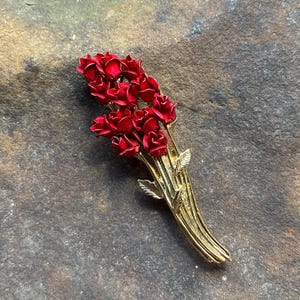 May include: A gold-tone metal brooch shaped like a bouquet of red roses. The piece features multiple small, deep red enameled rosebuds clustered at the top, with textured gold stems and leaves extending downward to form the base of the floral arrangement.