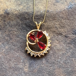 May include: A gold-colored pendant necklace with a circular design. The pendant features a red, flower-like arrangement of gemstones in the center, surrounded by a ring of smaller, clear gemstones. The necklace chain is also gold-colored.