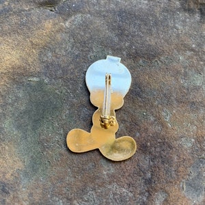 Pu&ograve; includere: Spilla in metallo color oro raffigurante un personaggio dei cartoni animati con una grande testa e piedi. La spilla ha una parte superiore color argento e un corpo color oro. La spilla &egrave; attaccata a una chiusura color oro.