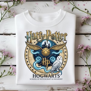 May include: White sweatshirt featuring a circular graphic with the words "Harry Potter" in gold lettering. The design includes a golden snitch, a snowy owl, and the Hogwarts crest. The text "Hogwarts School of Witchcraft & Wizardry" is also present.