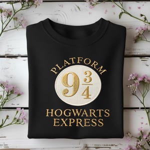 Puede incluir: Una sudadera negra de cuello redondo con un parche circular bordado en el centro. El parche es de color crema con texto dorado que dice Platform 9 3/4 y Hogwarts Express, con el número 9 3/4 mostrado de forma prominente en color dorado.