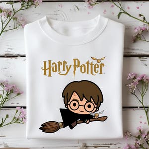 May include: A white crew neck t-shirt featuring a gold Harry Potter logo at the top and a cartoon illustration of Harry Potter with brown hair and round glasses riding a brown broomstick while holding a small golden snitch in the center.