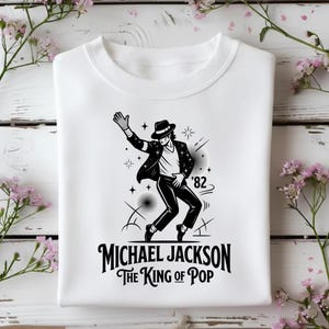 Può includere: Felpa bianca con grafica in bianco e nero di Michael Jackson in una posa iconica. La grafica include il testo "MICHAEL JACKSON THE KING OF POP" e l'anno '82.