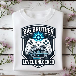 May include: White sweatshirt featuring a graphic with the text "BIG BROTHER" above a video game controller design. Below the controller is the text "LEVEL UNLOCKED". The design is in shades of blue and white.