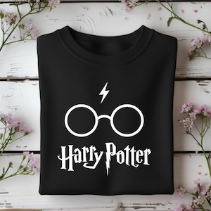 Puede incluir: Una sudadera negra de cuello redondo con un estampado gráfico blanco de gafas redondas y un rayo sobre el texto Harry Potter, escrito en una fuente estilizada con un detalle de rayo en la letra P.
