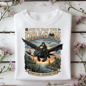 May include: White sweatshirt featuring a colorful graphic from "How to Train Your Dragon." The design shows a person riding a black dragon over a coastal landscape. Text includes "How to Train Your Dragon" and "Strike Class - Unstoppable Bond."