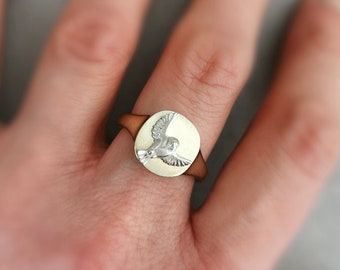 May include: A signet ring featuring a circular face with a polished gold-tone finish and a raised silver-tone bird emblem in flight. The band of the ring is a solid gold-tone metal, creating a two-tone appearance on the finger.