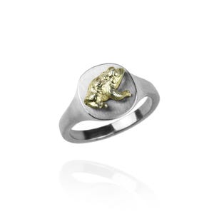 Puede incluir: Un anillo de sello de plata con un diseño de rana de oro en la parte superior.