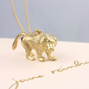 Op de afbeelding: Een gouden ketting met een gedetailleerde baviaan hanger. De baviaan loopt naar voren met zijn hoofd naar beneden gekanteld. De baviaan heeft een rode edelsteen op zijn achterwerk.