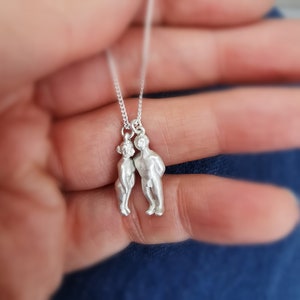 Puede incluir: Un collar de plata con dos pequeñas figuras de plata de personas de pie una al lado de la otra. Las figuras se miran y parecen estar cogidas de la mano.