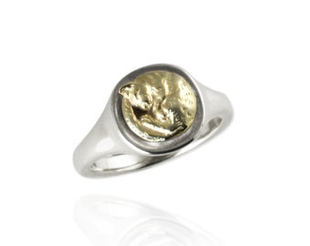 May include: A silver signet ring featuring a circular face with a raised, gold-toned relief of a curled-up cat. The band is polished silver, and the setting surrounding the gold cat figure is darkened to provide contrast to the metallic design.