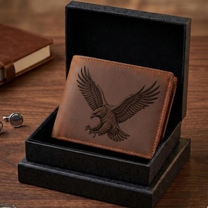 May include: A brown leather wallet with an engraved eagle design, presented in a black gift box. The wallet is open, showcasing the detailed artwork. A brown leather notebook and silver cufflinks are also visible.