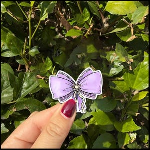 May include: A sticker featuring a lavender bow with a yellow center and dangling ribbons. The bow has a detailed design with a decorative border. The sticker is held against a backdrop of green leaves.