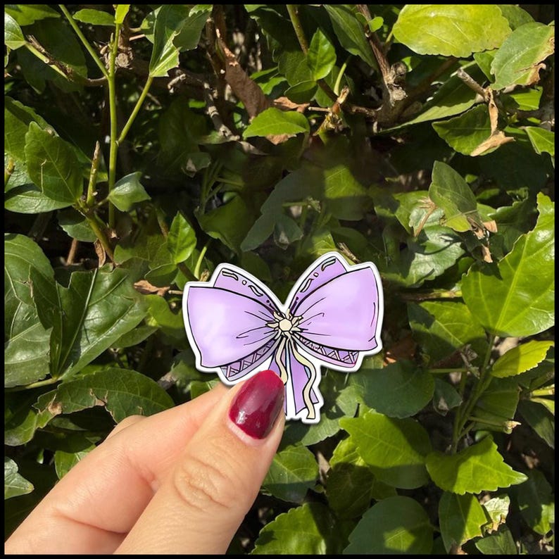 May include: A sticker featuring a lavender bow with a yellow center and dangling ribbons. The bow has a detailed design with a decorative border. The sticker is held against a backdrop of green leaves.