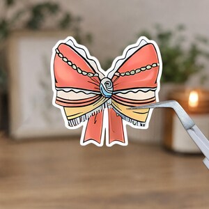 May include: A sticker featuring a detailed illustration of a bow. The bow is primarily coral-colored with white lace trim, a blue and white center, and yellow and black striped accents. A pair of tweezers holds the sticker.