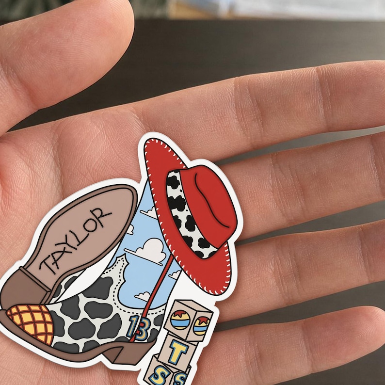 Puede incluir: Una pegatina troquelada que muestra una bota de vaquero con estampado de vaca, un fondo de cielo azul con nubes y el n&uacute;mero 13. Un sombrero de vaquero rojo con una banda de estampado de vaca se sit&uacute;a sobre la bota. La palabra TAYLOR est&aacute; escrita.